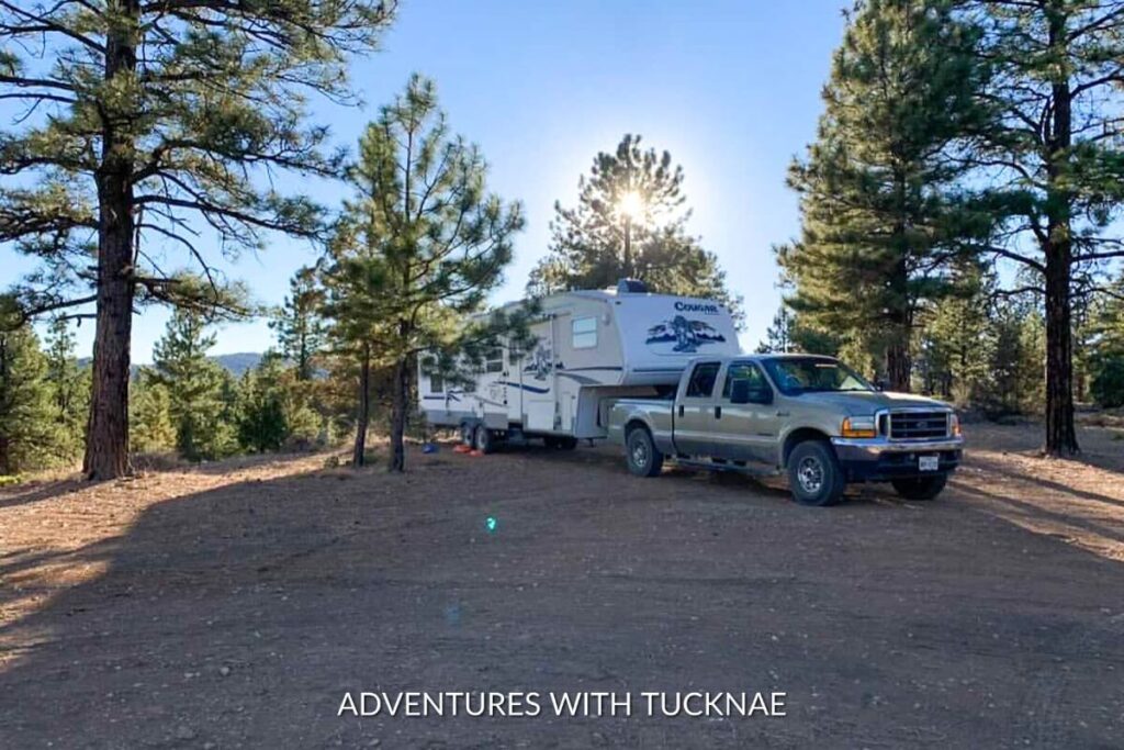 Fifth-wheel RV and pickup truck set up in a forest clearing, with tall pines around and the sun setting behind, offering a secluded boondocking experience.