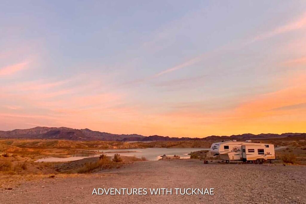 A modern travel trailer sits by a tranquil lake at dusk, with soft pink skies reflecting the calmness of boondocking in solitude.