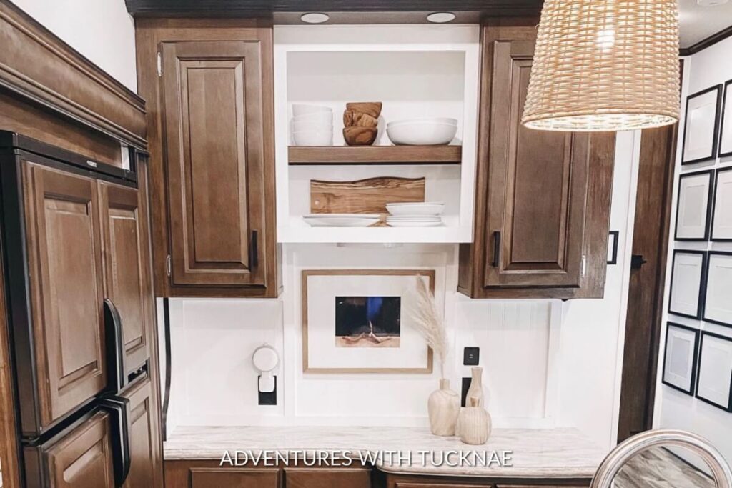 Modern RV kitchen with brown wooden cabinets, white open shelving displaying clean dishes, and a woven pendant light.