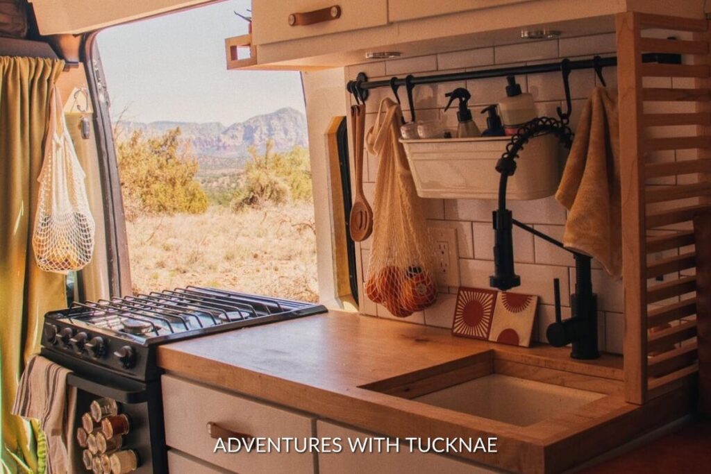 Rustic RV kitchen setup in a van with a scenic desert view, featuring a gas stove, hanging fruits, and wooden cabinetry.