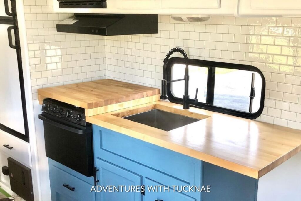 Compact RV kitchen corner featuring a black farmhouse sink, blue cabinets, and butcher block counters against a subway tile backsplash.