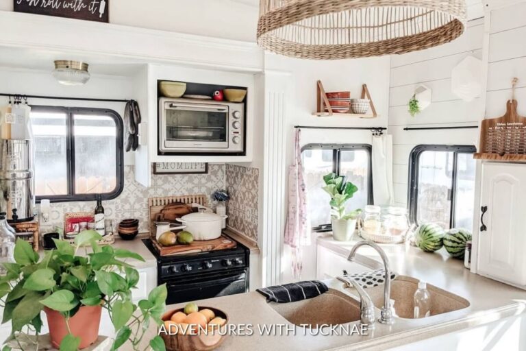 Bright and airy RV kitchen with natural light, featuring a woven hanging light fixture, white cabinetry, a microwave, and a lively plant on the counter. Decorative touches include a sign, a patterned backsplash, and neatly arranged kitchen utensils.