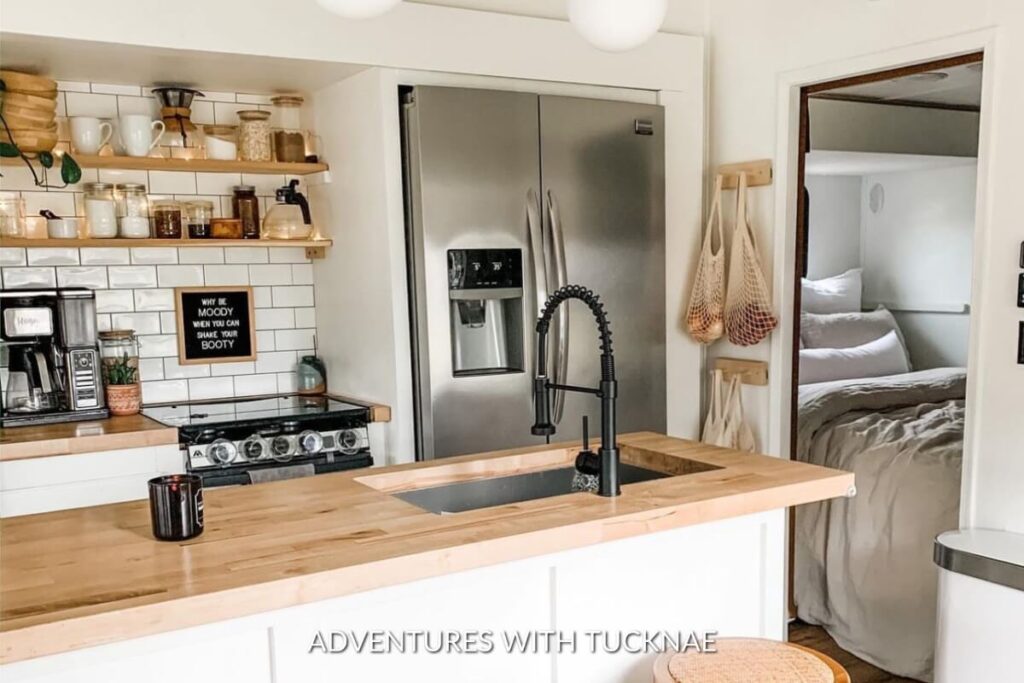 Bright RV kitchen with a witty wall sign, floating wooden shelves filled with glass jars, and a stainless steel fridge.