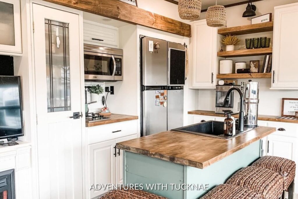 White and wood-themed RV kitchen with a central island, stainless steel appliances, and rustic hanging lights.