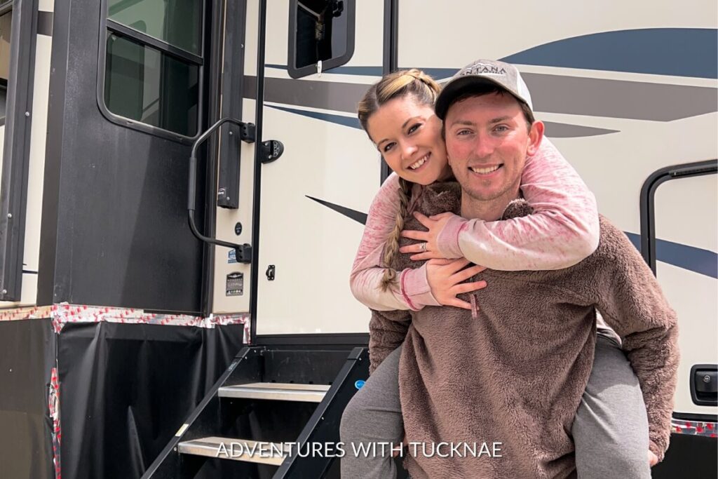 Janae wraps her arms around Tucker from behind, both smiling brightly, standing in front of the open door of their RV, suggesting a sense of adventure and companionship.