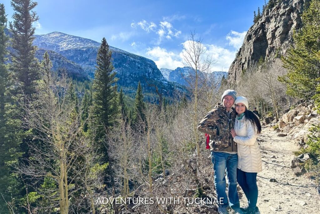 Tucker and Janae stand smiling on a rocky mountain trail, clad in winter gear, with majestic snow-covered peaks and evergreen trees under a clear blue sky.