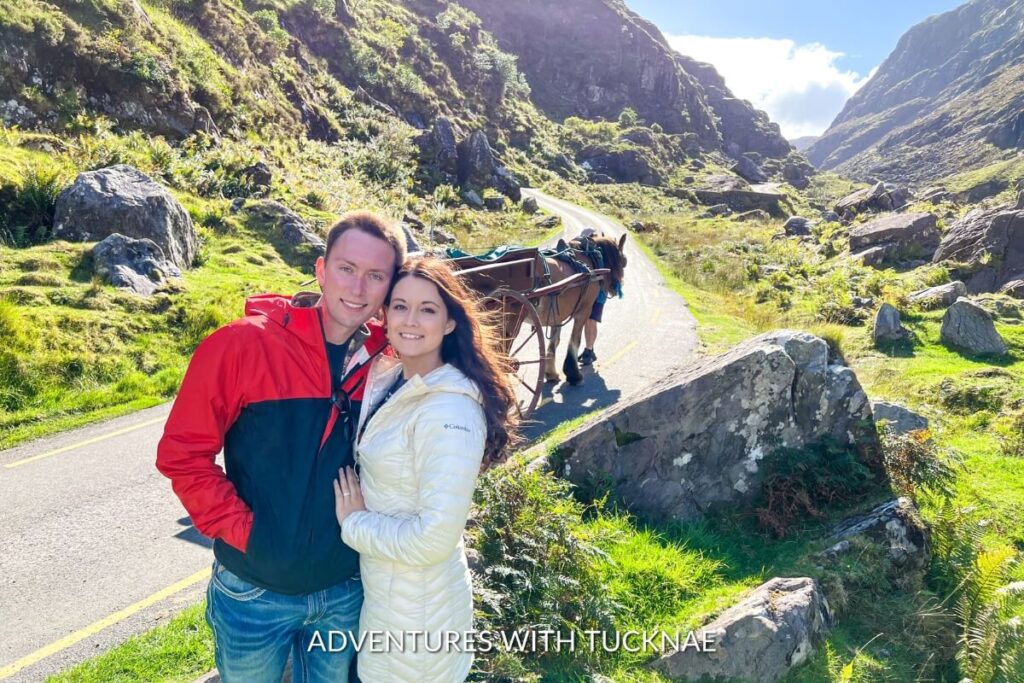Tucker and Janae share a close moment on a serene mountain path, with a traditional horse-drawn carriage in the background amidst the lush green hills of the Gap of Dunloe in Ireland.