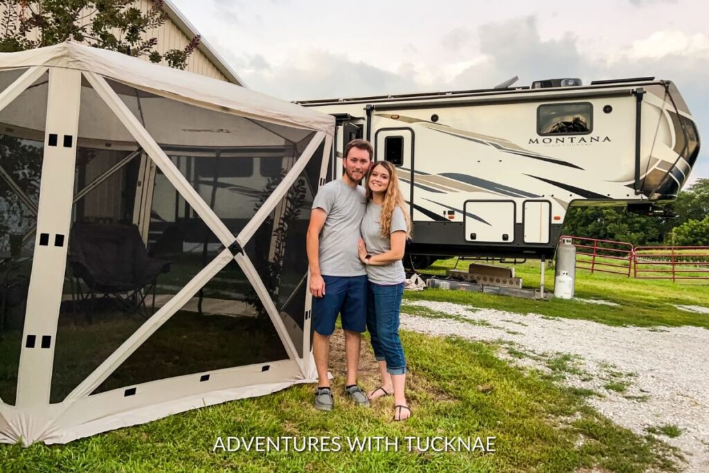 A smiling couple standing in front of their fifth-wheel RV with their Lippert pop-up gazebo.