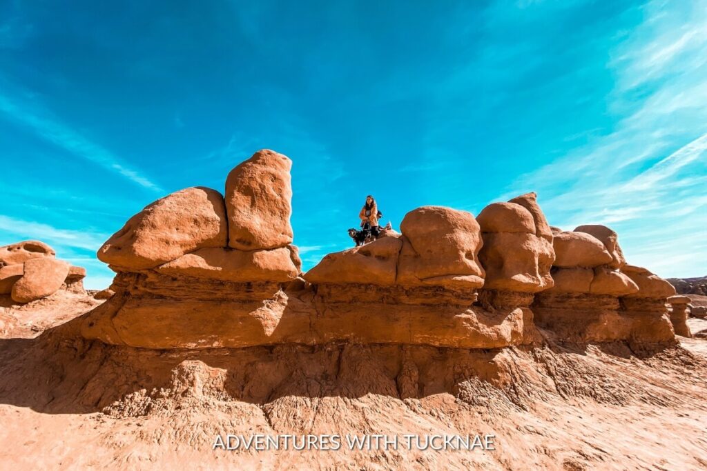 A person poses with a dog on a unique rock formation resembling goblin-like sculptures under the bright blue sky at Goblin Valley State Park, a whimsical and instagrammable destination in Utah.