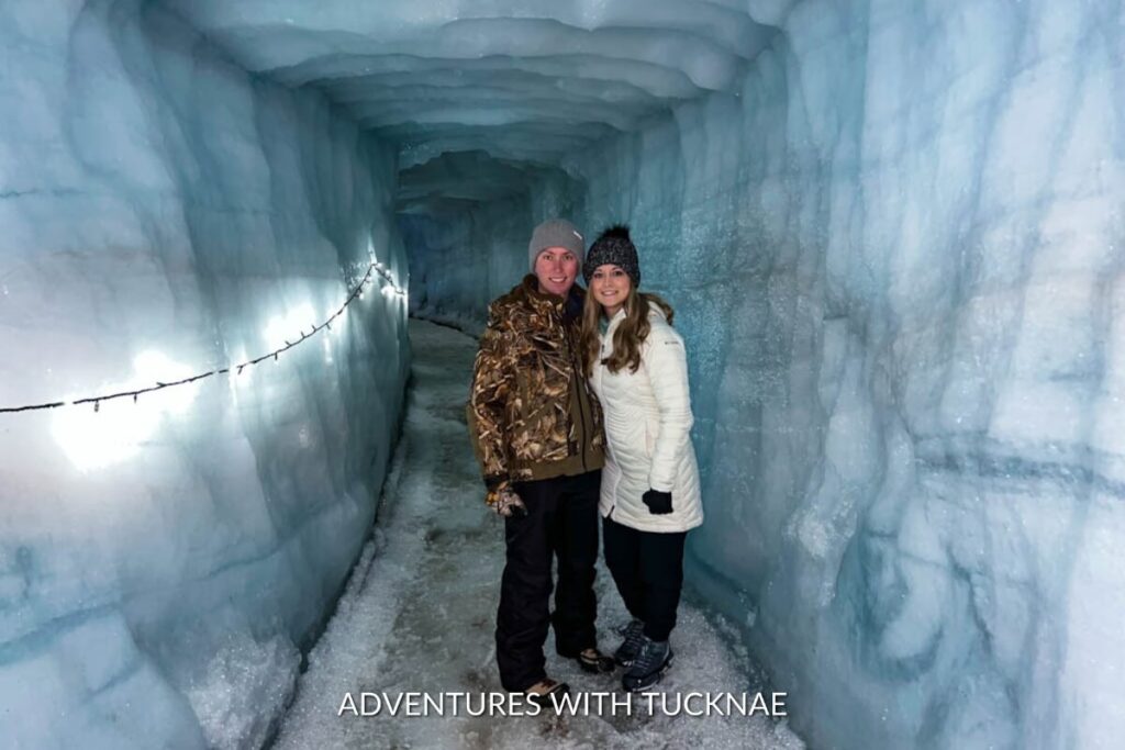 Tucker & Janae from Adventures With TuckNae exploring the icy depths of Into the Glacier, walking through a lit ice tunnel with rugged, frozen walls.