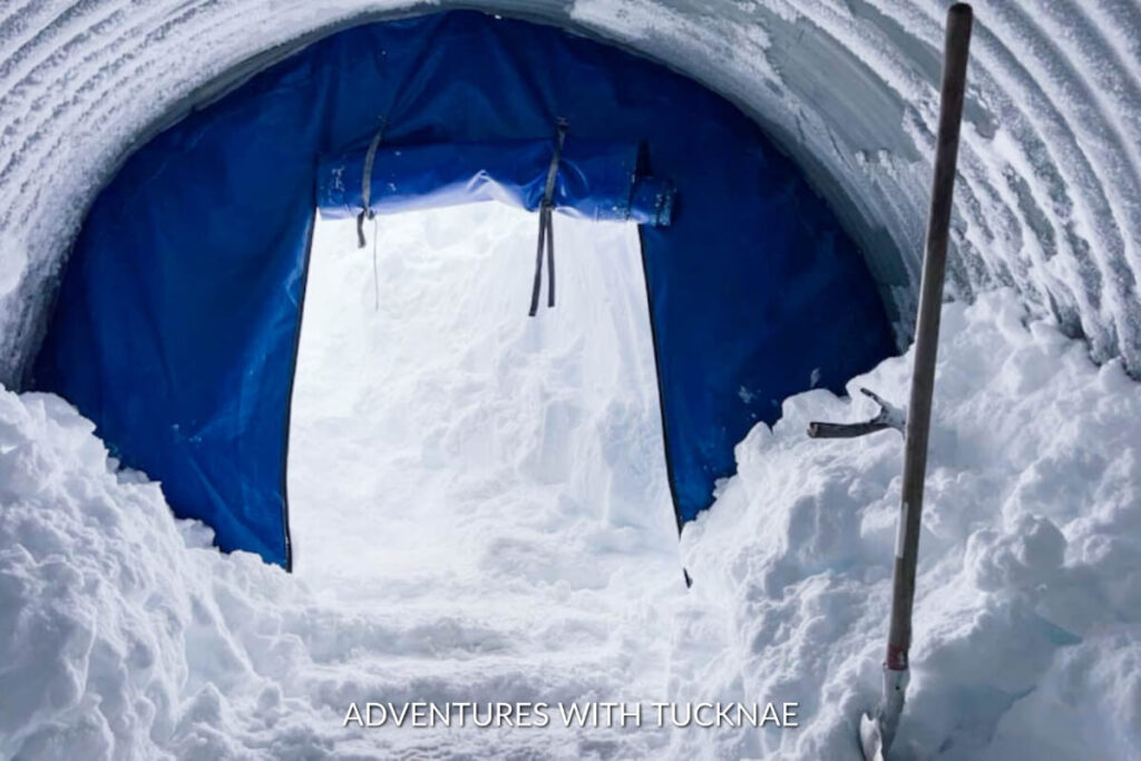 Entrance of the Into the Glacier tunnel draped with a blue tarp, framed by snow-covered walls and a shovel planted in the snow.