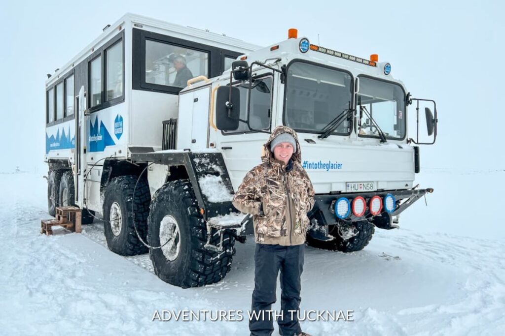 Adventurous traveler standing proudly in front of the Into the Glacier ice explorer truck on a snowy terrain, under a cloudy sky.