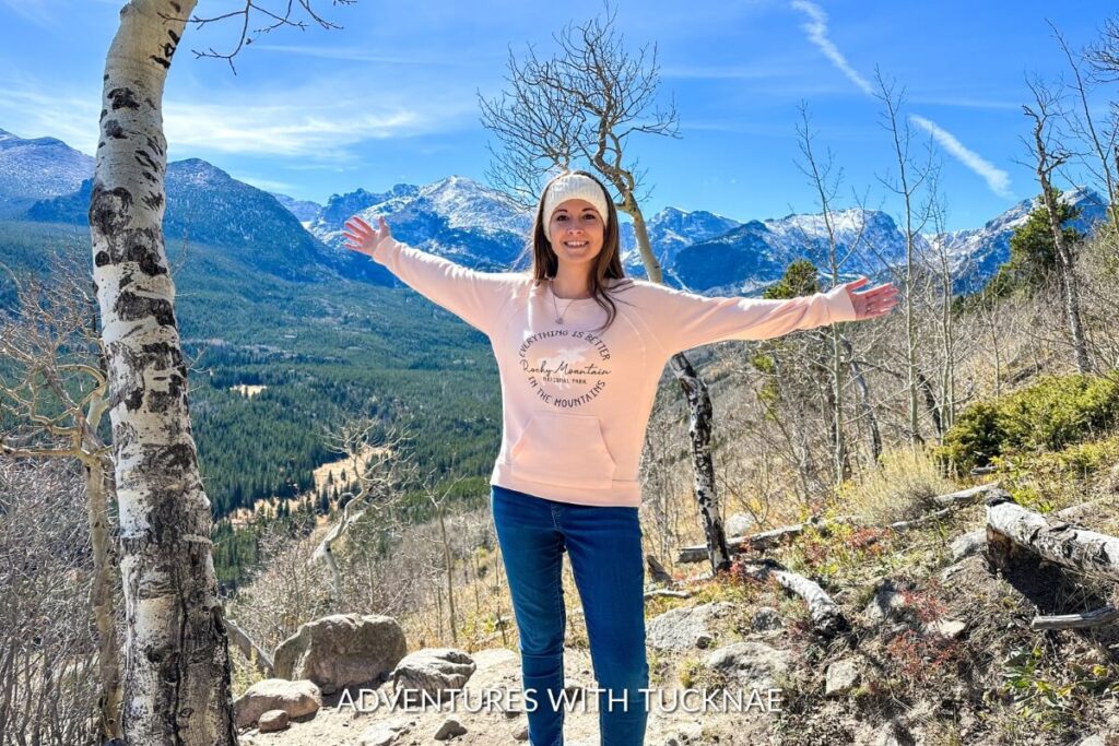 Janae stands with her arms outstretched, embracing the grandeur of the Rocky Mountains, with snow-capped peaks and clear skies in the background.
