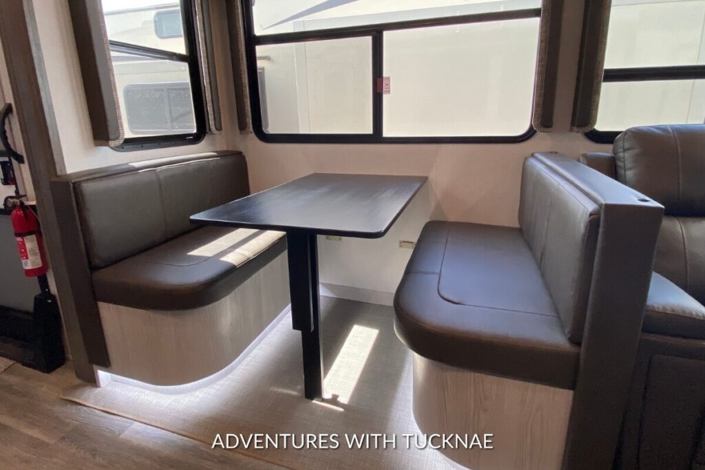 RV dining area with a brown booth-style table and adjacent leather seating in a Keystone Montana High Country RV.
