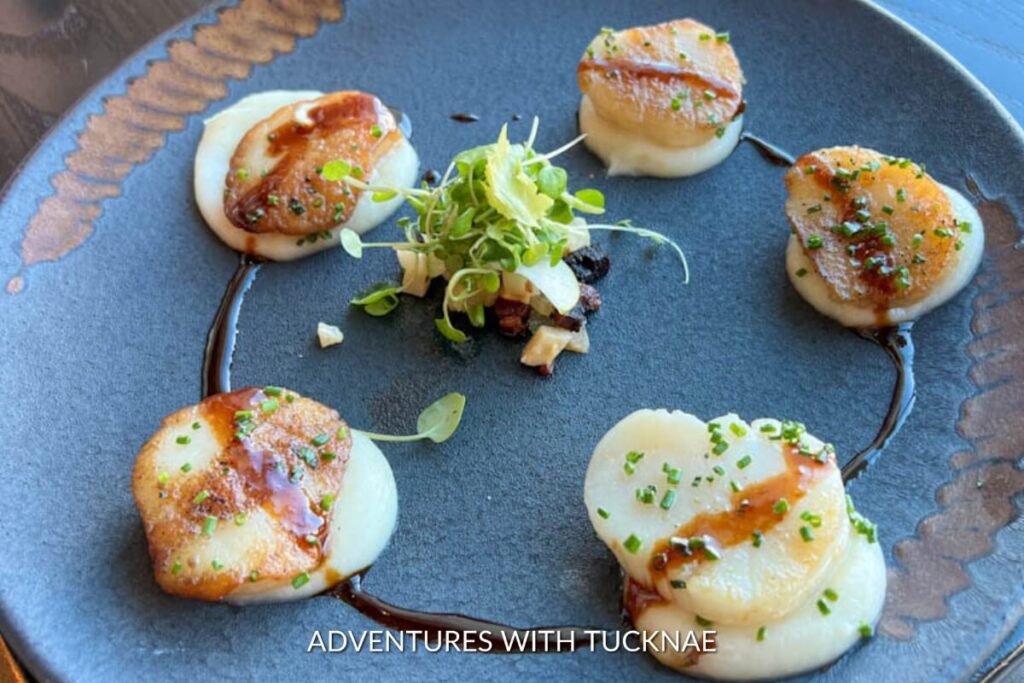 Pan-seared scallops with golden crust arranged on a slate plate, garnished with fresh greens and a decorative balsamic reduction, presented at a Las Vegas culinary adventure.