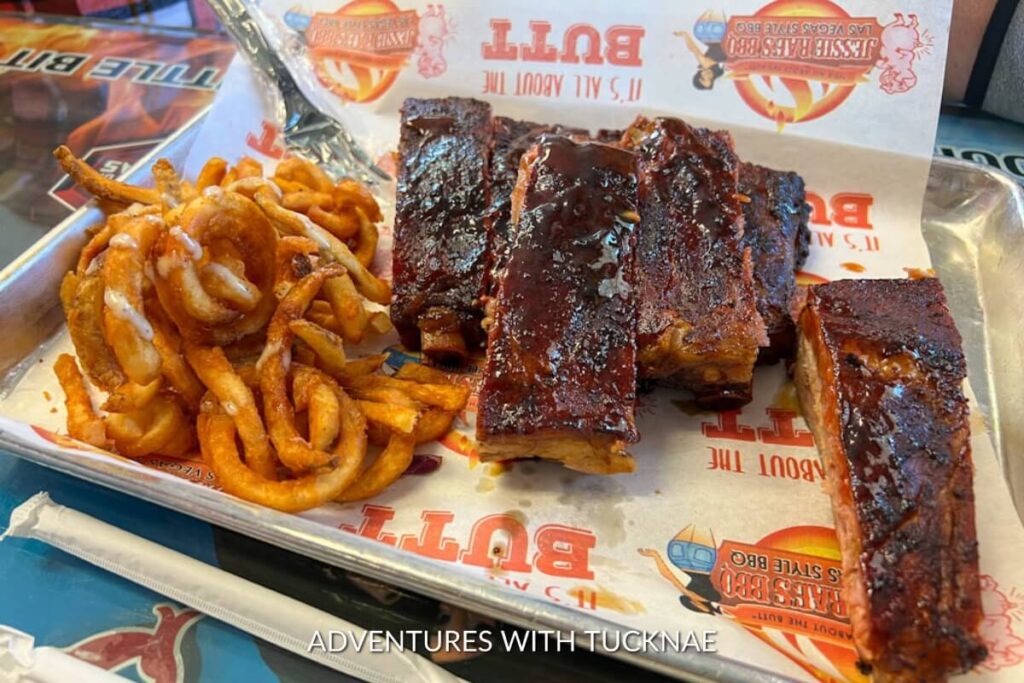 Succulent smoked ribs coated in a glossy BBQ sauce, paired with seasoned curly fries, presented on a paper-lined metal tray for a Las Vegas-style feast.