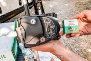 A close-up of a hand holding a Latch.it wireless key fob next to its packaging, with an RV electronic keypad lock in the background