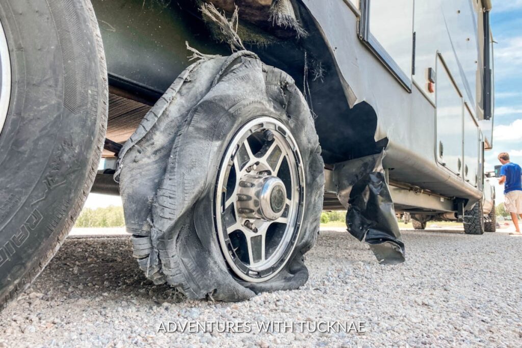 A damaged RV tire with extensive tread separation and a blown sidewall, with someone inspecting the damage, indicating a roadside issue