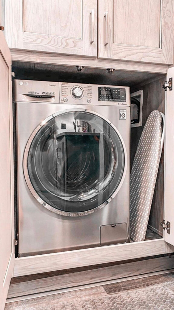 A sleek LG washing machine positioned next to a polka-dotted ironing board inside a wooden cabinet, exemplifying an efficient RV laundry setup.