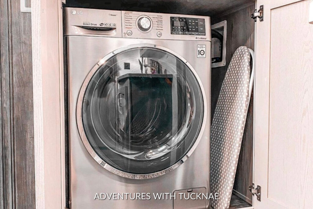 A sleek LG washing machine positioned next to a polka-dotted ironing board inside a wooden cabinet, exemplifying an efficient RV laundry setup.