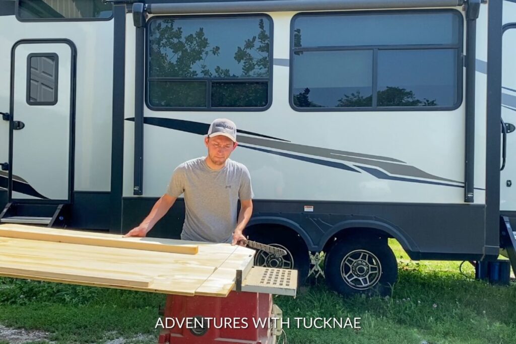 Outdoor view of a man preparing wooden planks for an RV project, with the RV in the background, showcasing the mobile lifestyle.