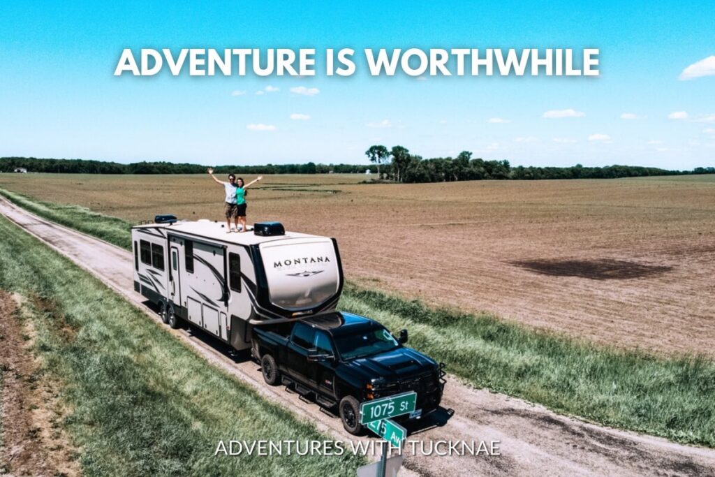 Aerial view of two people standing on top of an RV towed by a pickup truck on a rural road with the quote 'Adventure is worthwhile.'