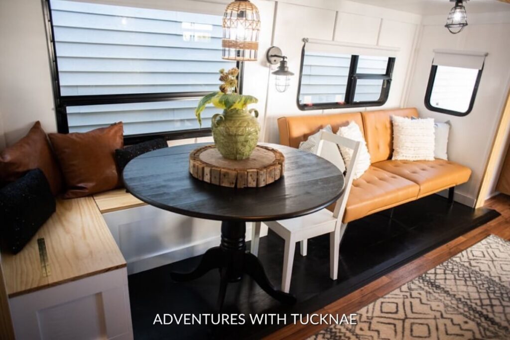 A round black table with a rustic wooden centerpiece, paired with a caramel-colored sofa bench in a well-lit RV interior.