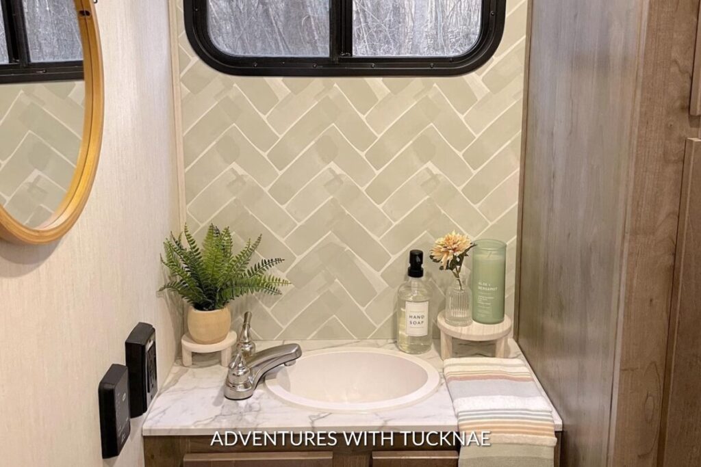 Elegant RV bathroom with a herringbone-patterned wallpaper in soft green, enhancing the space alongside a white countertop and a potted fern decoration.