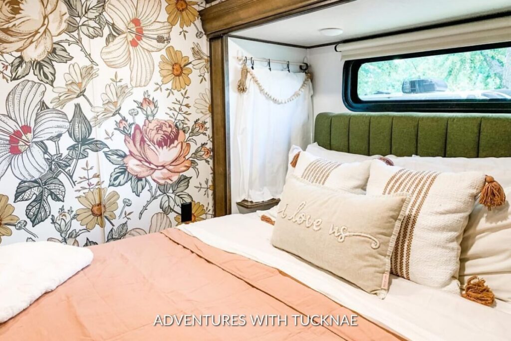 RV bedroom boasting a large floral wallpaper with bold botanical prints, complemented by 'olive us' embroidered pillows and a green upholstered headboard.