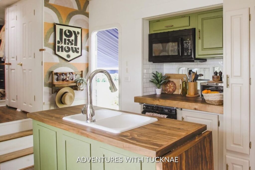 Eclectic RV kitchen area featuring a vibrant retro wallpaper with a 'Joy is Now' banner, contrasting with green cabinets and white subway tiles.