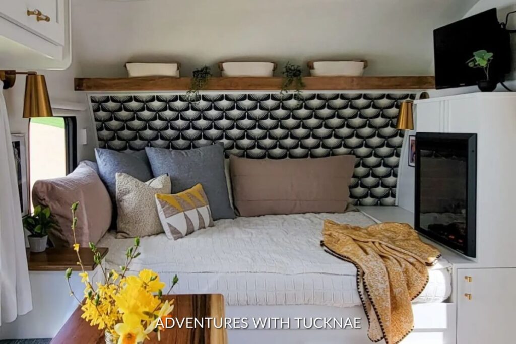 Stylish RV sleeping area highlighted by a bold black and white scallop-patterned wallpaper, complemented with soft pastel cushions and a wooden shelf above.