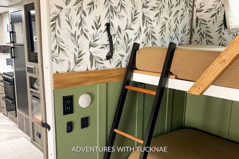 Smartly designed RV interior corner with a refreshing leaf-patterned wallpaper, a green cushioned bench seat, and a wooden ladder leading to an overhead nook, highlighting space efficiency and style.