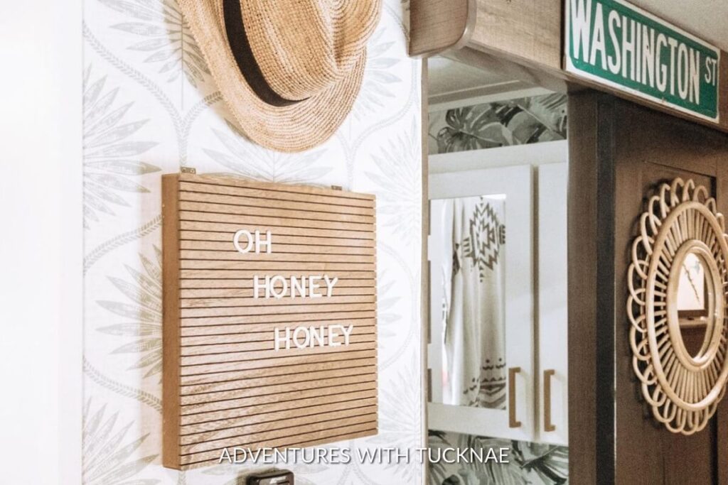 Quaint RV corner with a playful letter board against a leaf-patterned wallpaper, accompanied by a woven hat and a cozy blanket.