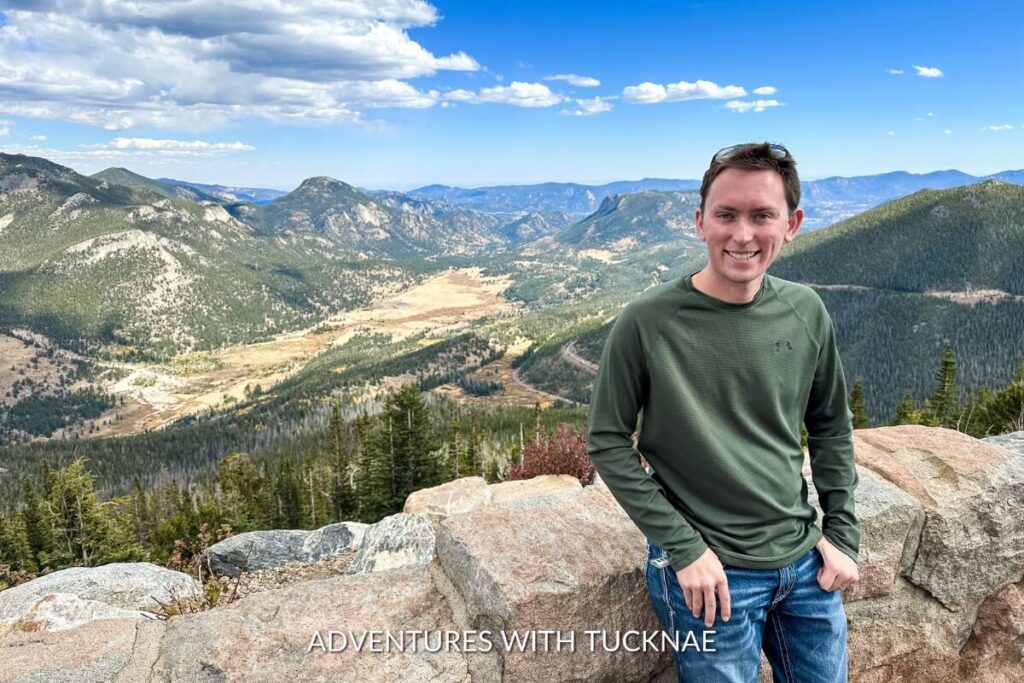 Tucker, casually dressed in a green long-sleeve shirt and jeans, poses with a gentle smile against a panoramic view of rolling forested hills and a wide mountain valley.