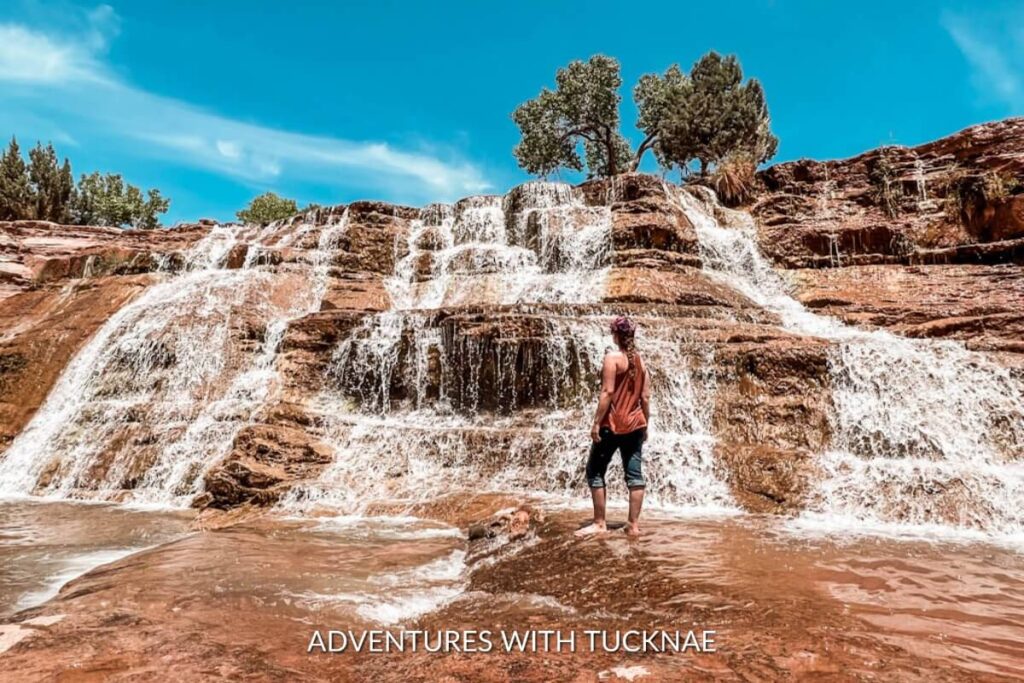 A person stands admiring Toquerville Falls, a cascade of clear waters over terracotta-colored rocks under a blue sky, in Southern Utah.