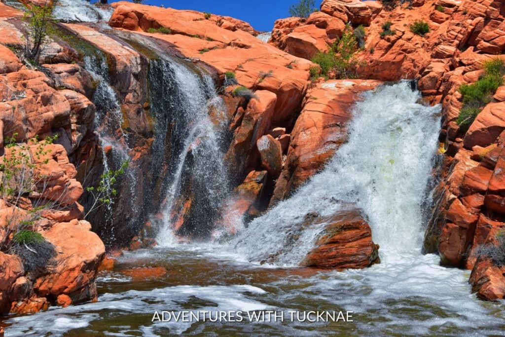 Vibrant red rocks surround the flowing Gunlock Falls, where water splashes into a clear pool below under the bright Utah sky.