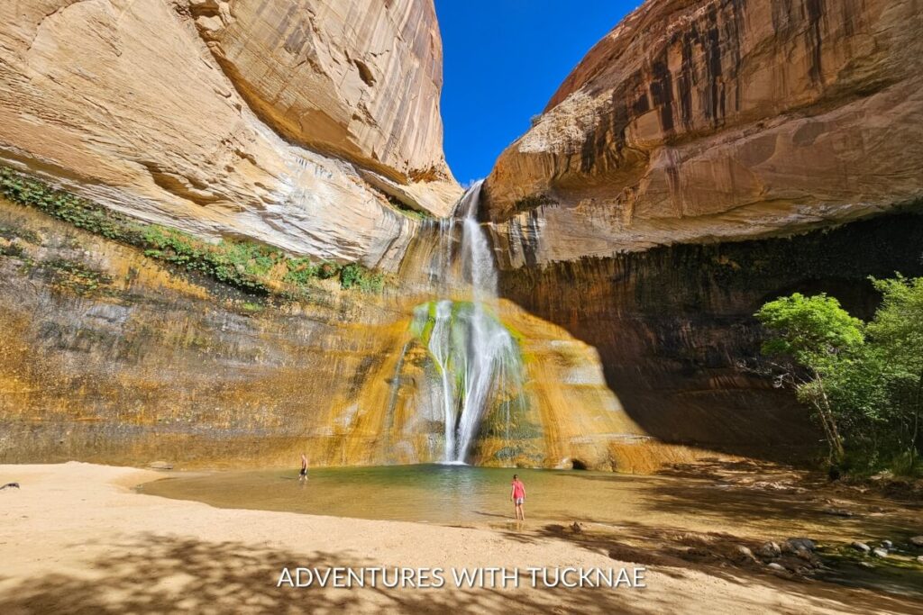 Visitors at Lower Calf Creek Falls enjoy the view of water plummeting down a large, ochre cliff face into a sandy basin in Southern Utah.