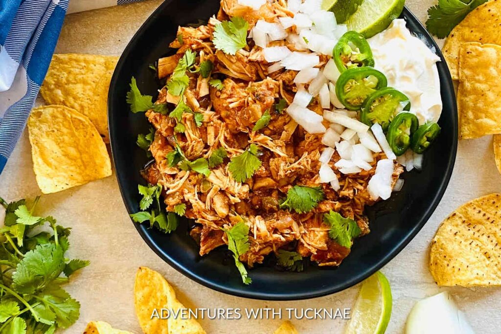 Instant Pot Salsa Chicken: A black bowl filled with Instant Pot salsa chicken topped with fresh cilantro, diced onions, and jalapeños, surrounded by tortilla chips.