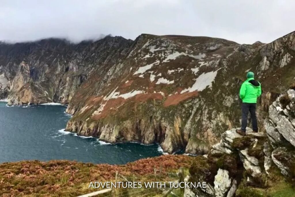 A person in a bright green jacket stands on the cliff edge at Slieve League, one of the most Instagram-worthy spots in Ireland, with dramatic coastal views stretching into the horizon.