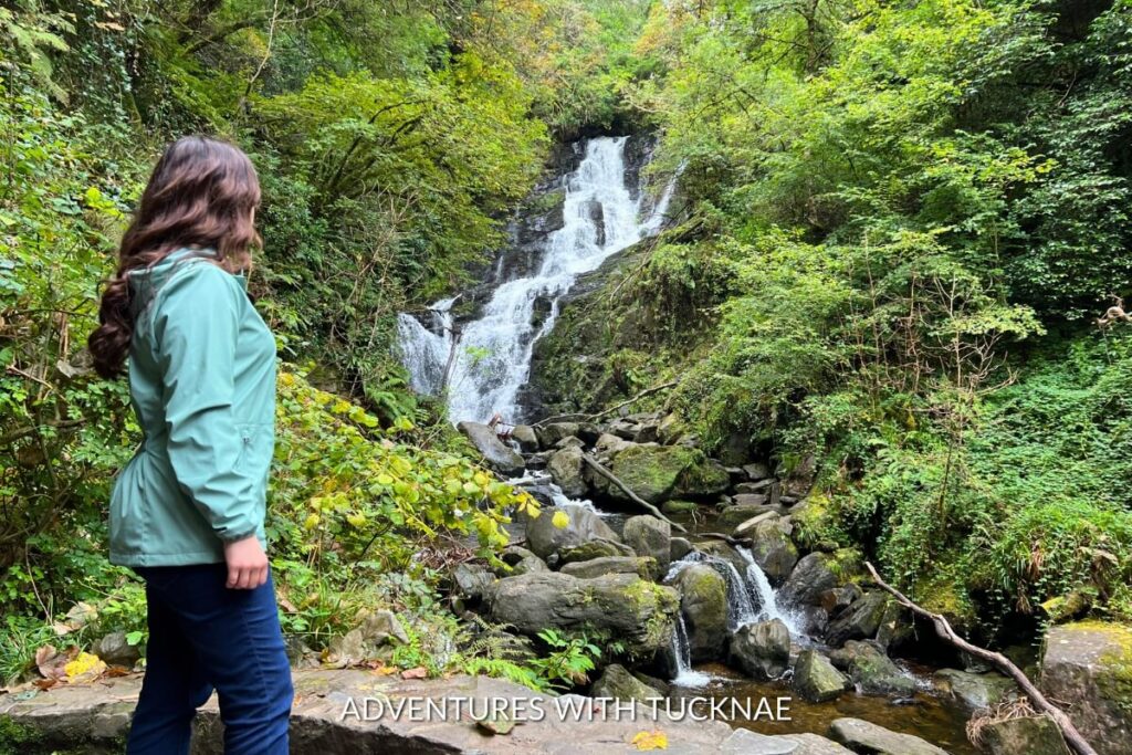 A woman gazes at Torc Waterfall nestled within lush greenery, a tranquil and Instagrammable location in Ireland's Killarney National Park.