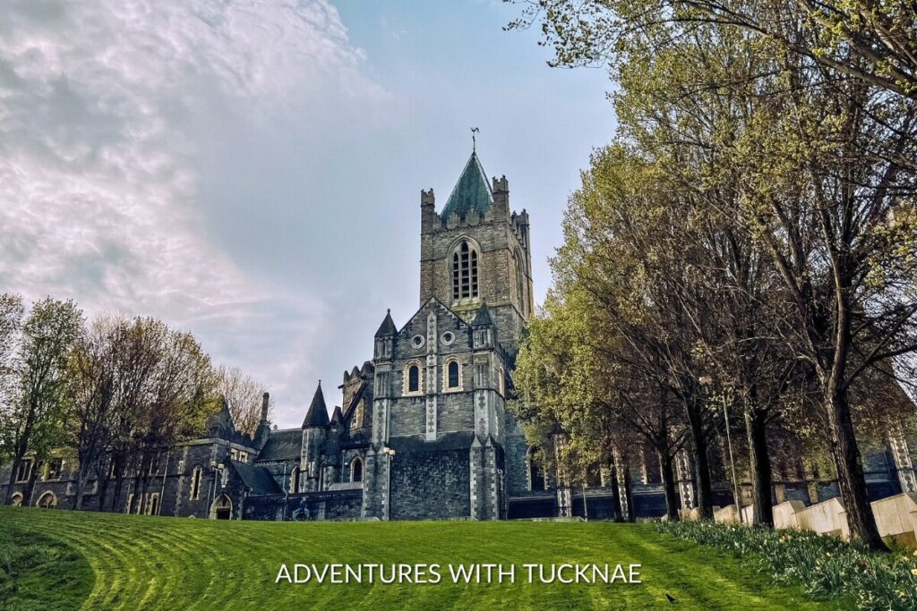 The majestic Christ Church Cathedral under a cloudy sky, surrounded by lush greenery, stands as a testament to Dublin's architectural splendor and Instagrammable heritage.