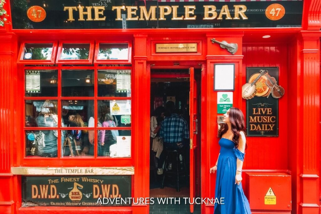 Vibrant red exterior of the Temple Bar, a bustling and Instagrammable hotspot in Dublin, with a woman in a blue dress admiring the lively atmosphere.