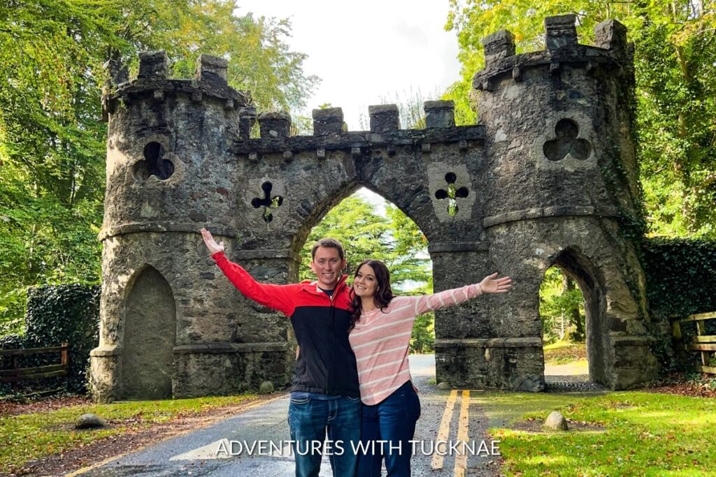 A couple with outstretched arms stands in front of the whimsical gatehouse at Tollymore Forest Park, adding charm to a weekend break in Ireland.