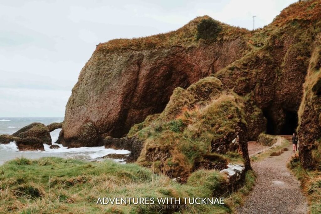 A rugged cave entrance at the base of a lush cliff in Cushendall, inviting exploration for those on a weekend break in Ireland.
