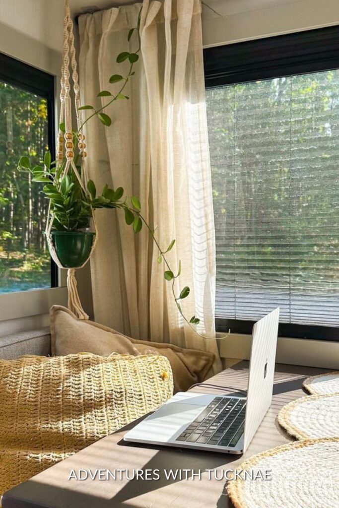 A hanging potted plant cascading over a macramé hanger beside a sunlit RV window, with a cozy seating area and open laptop, creating a perfect nook for work or relaxation among nature.