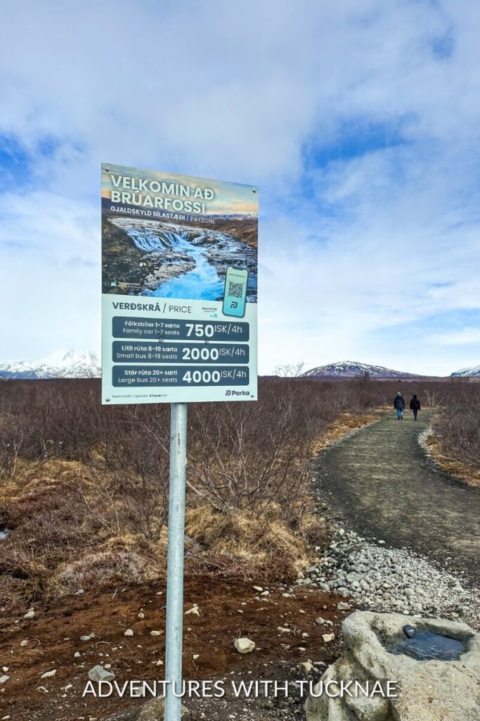 A trail leading to Brúarfoss waterfall in Iceland with a sign listing parking prices; the snowy mountain and barren landscape stretch in the background.
