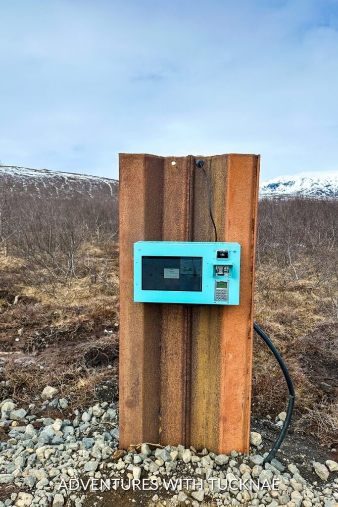 A rustic parking meter station on a trail in Iceland with a rusty orange panel and a teal digital interface, surrounded by a rocky landscape with sparse vegetation.