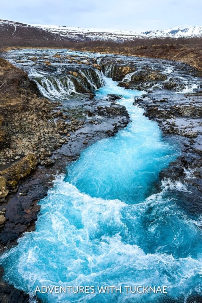 The vibrant blue waters of Brúarfoss waterfall in Iceland cascading through rugged terrain, with snow-covered mountains in the distance.