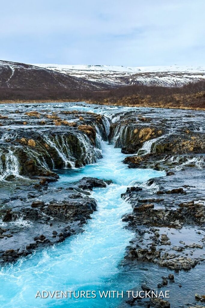 Landscape view of Brúarfoss waterfall flowing vigorously through a rocky riverbed, highlighted by its distinctive blue color, with a barren, snow-dusted landscape around.