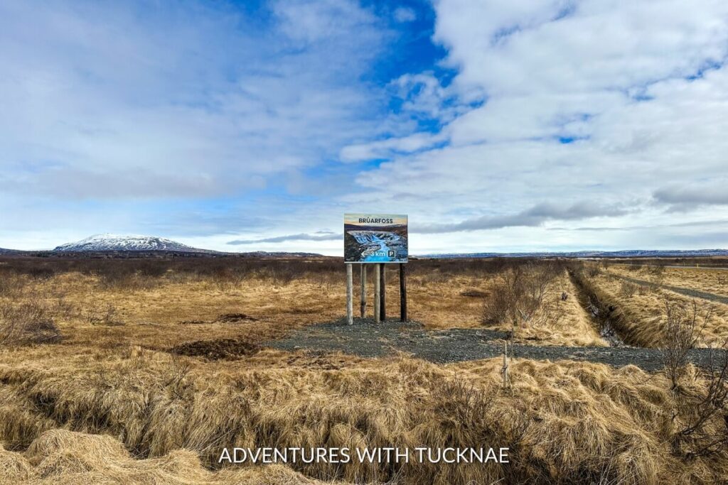 Signage at the beginning of the road to Brúarfoss waterfall, displaying a picture of the blue waterfall and indicating a 3 km driving distance, set against a backdrop of flat, grassy terrain and distant snow-capped mountains under a cloudy sky.